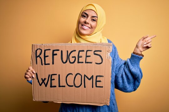 Beautiful arab woman wearing islamic hijab holding banner with refugees welcome message very happy pointing with hand and finger to the side