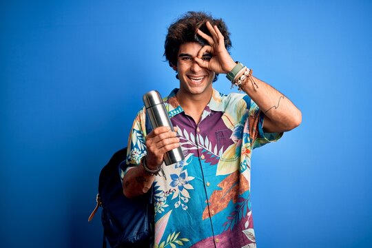 Young tourist man wearing backpack holding thermo over isolated blue background with happy face smiling doing ok sign with hand on eye looking through fingers