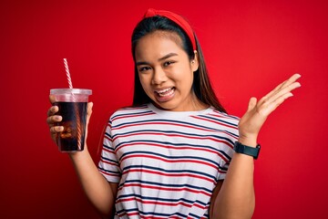 Young asian girl drinking cola fizzy refreshment using straw over isolated red background very happy and excited, winner expression celebrating victory screaming with big smile and raised hands