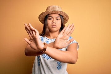 Young beautiful asian girl wearing casual t-shirt and hat standing over yellow background Rejection expression crossing arms doing negative sign, angry face