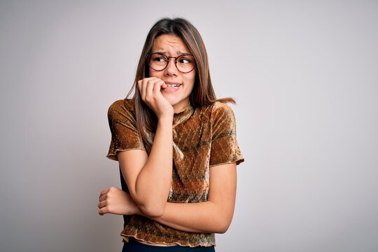 Young beautiful brunette girl wearing casual t-shirt and glasses over isolated white background looking stressed and nervous with hands on mouth biting nails. Anxiety problem.