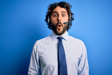 Young handsome call center agent man with beard working using headset over blue background afraid and shocked with surprise expression, fear and excited face.