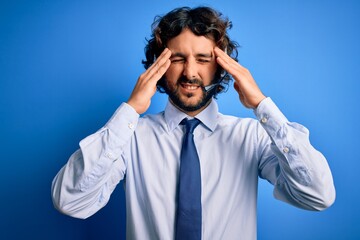 Young handsome call center agent man with beard working using headset over blue background suffering from headache desperate and stressed because pain and migraine. Hands on head.