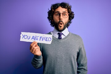 Handsome business man with beard holding you are fired message over purple background scared in shock with a surprise face, afraid and excited with fear expression