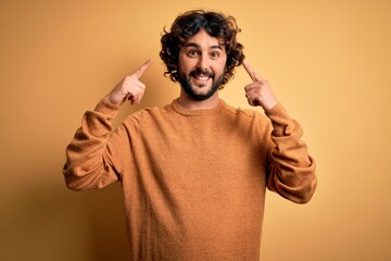 Young handsome man with beard wearing casual sweater standing over yellow background smiling pointing to head with both hands finger, great idea or thought, good memory