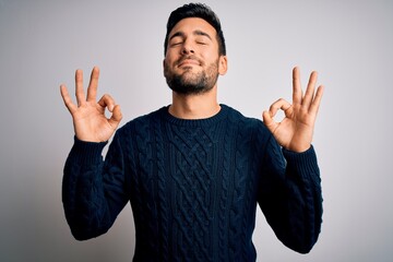 Naklejka premium Young handsome man with beard wearing casual sweater standing over white background relax and smiling with eyes closed doing meditation gesture with fingers. Yoga concept.