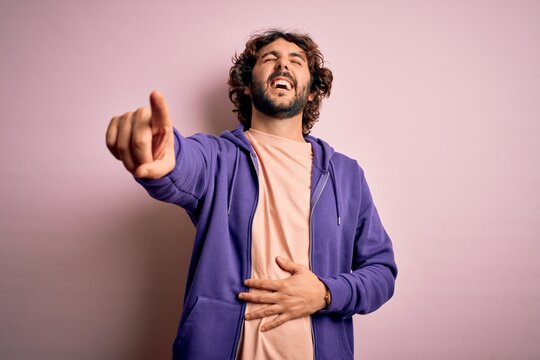 Young handsome sporty man with beard wearing casual sweatshirt over pink background laughing at you, pointing finger to the camera with hand over body, shame expression