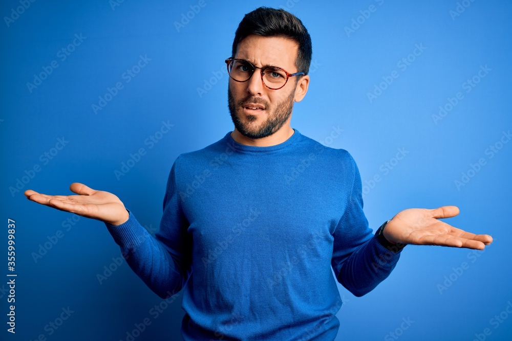 Wall mural young handsome man with beard wearing casual sweater and glasses over blue background clueless and c