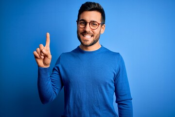 Young handsome man with beard wearing casual sweater and glasses over blue background showing and...