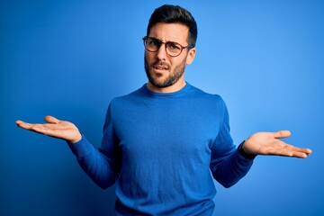 Young handsome man with beard wearing casual sweater and glasses over blue background clueless and confused with open arms, no idea concept.