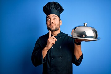 Young cooker man with beard wearing uniform holding tray with dome over blue background serious face thinking about question, very confused idea