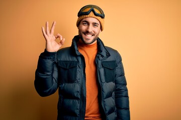 Young handsome skier man with beard wearing snow sportswear and ski goggles smiling positive doing ok sign with hand and fingers. Successful expression.