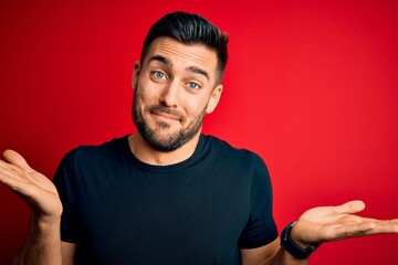 Young handsome man wearing casual black t-shirt standing over isolated red background clueless and confused expression with arms and hands raised. Doubt concept.