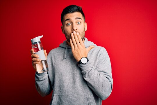 Young handsome sportsman drinking bottle of water to refeshment over red background cover mouth with hand shocked with shame for mistake, expression of fear, scared in silence, secret concept