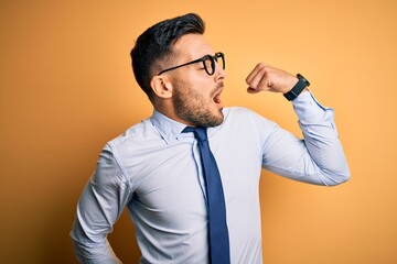 Young handsome businessman wearing tie and glasses standing over yellow background stretching back, tired and relaxed, sleepy and yawning for early morning