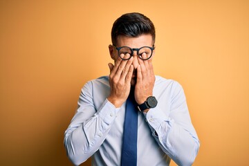 Young handsome businessman wearing tie and glasses standing over yellow background rubbing eyes for...