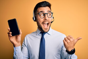 Business operator man with customer service headset from call center showing smartphone screen pointing and showing with thumb up to the side with happy face smiling
