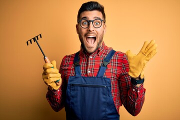 Young gardener man wearing working apron using gloves and tool over yellow background very happy...