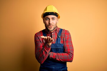 Young builder man wearing construction uniform and safety helmet over yellow isolated background looking at the camera blowing a kiss with hand on air being lovely and sexy. Love expression.