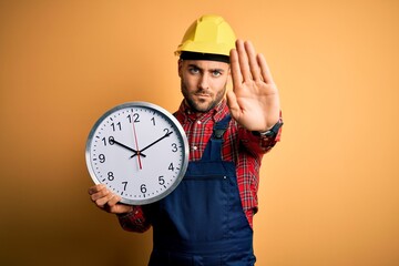 Young builder man wearing safety helmet holding big clock over yellow background with open hand...