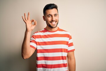 Fototapeta premium Young handsome man wearing casual striped t-shirt standing over isolated white background smiling positive doing ok sign with hand and fingers. Successful expression.
