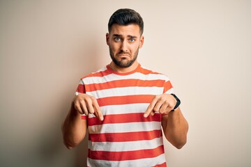 Young handsome man wearing casual striped t-shirt standing over isolated white background Pointing down looking sad and upset, indicating direction with fingers, unhappy and depressed.