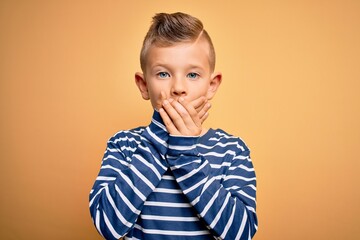 Young little caucasian kid with blue eyes wearing nautical striped shirt over yellow background shocked covering mouth with hands for mistake. Secret concept.