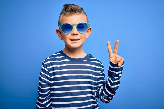 Young little caucasian kid with blue eyes standing wearing sunglasses over blue background smiling with happy face winking at the camera doing victory sign. Number two.