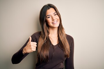 Young beautiful girl wearing casual sweater standing over isolated white background doing happy thumbs up gesture with hand. Approving expression looking at the camera showing success.
