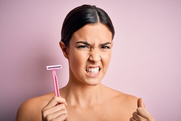 Young beautiful girl using shaver for depilation standing over isolated pink background annoyed and...