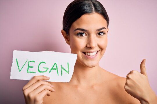 Young beautiful girl holding paper with vegan message over isolated pink background happy with big smile doing ok sign, thumb up with fingers, excellent sign