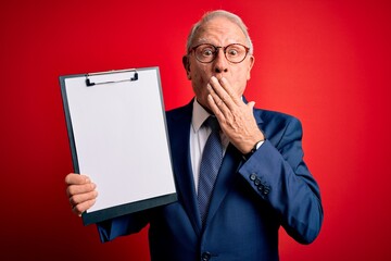 Senior grey haired business man holding clipboard over red background cover mouth with hand shocked with shame for mistake, expression of fear, scared in silence, secret concept