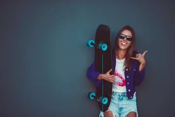 Abwaschbare Fototapete Young happy girl in sunglasses showing shaka with skateboard in front of grey wall. © Creative Cat Studio