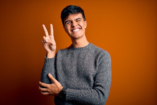 Young handsome man wearing casual sweater standing over isolated orange background smiling with happy face winking at the camera doing victory sign with fingers. Number two.