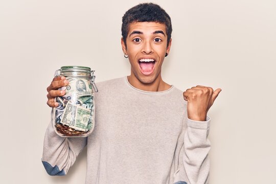 Young african amercian man holding jar with savings pointing thumb up to the side smiling happy with open mouth