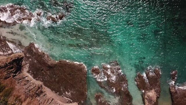 Exotic 4K Aerial Overhead of Cristal Clear War Beach with Dark Rocks and Waves. Beautiful Ocean Reflections of the Sun. Drone Movement Forward.