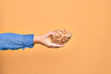 Obraz premium Beautiful hand of woman holding bowl with healthy salty peanuts over isolated yellow background