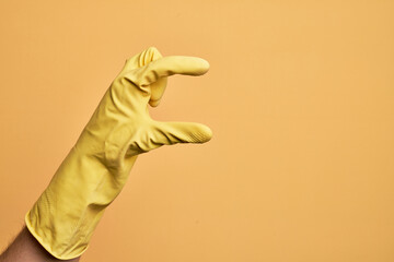 Hand of caucasian young man with cleaning glove over isolated yellow background picking and taking invisible thing, holding object with fingers showing space