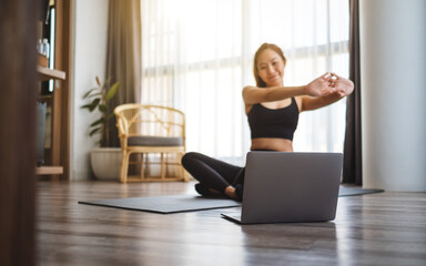 A beautiful young asian woman stretching arms while watching online workout tutorials on laptop at...