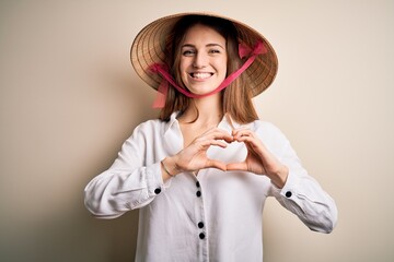 Young beautiful redhead woman wearing asian traditional conical hat over white background smiling in love showing heart symbol and shape with hands. Romantic concept.