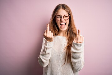 Young beautiful redhead woman wearing casual sweater and glasses over pink background Showing middle finger doing fuck you bad expression, provocation and rude attitude. Screaming excited