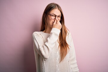 Young beautiful redhead woman wearing casual sweater and glasses over pink background smelling something stinky and disgusting, intolerable smell, holding breath with fingers on nose. Bad smell