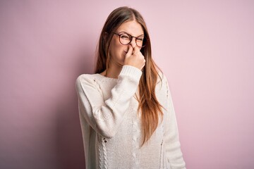 Young beautiful redhead woman wearing casual sweater and glasses over pink background smelling...