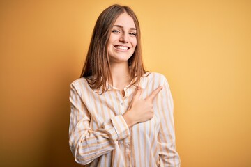 Young beautiful redhead woman wearing casual striped shirt over isolated yellow background cheerful with a smile of face pointing with hand and finger up to the side with happy and natural expression