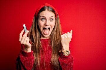 Young beautiful redhead woman holding USB memory over isolated red background screaming proud and celebrating victory and success very excited, cheering emotion
