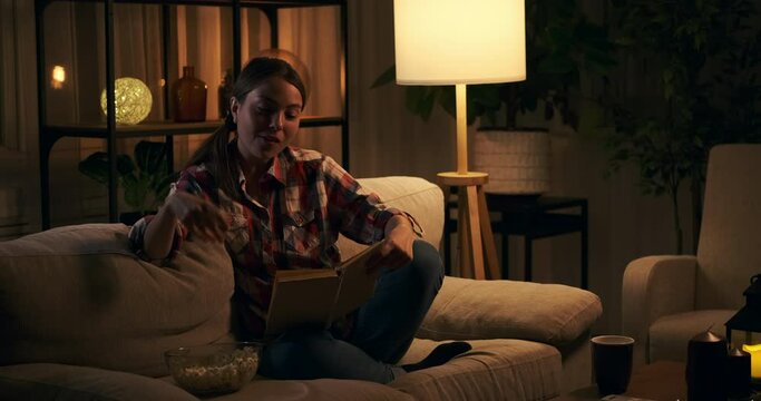Happy woman reading a book while eating popcorn on sofa at night