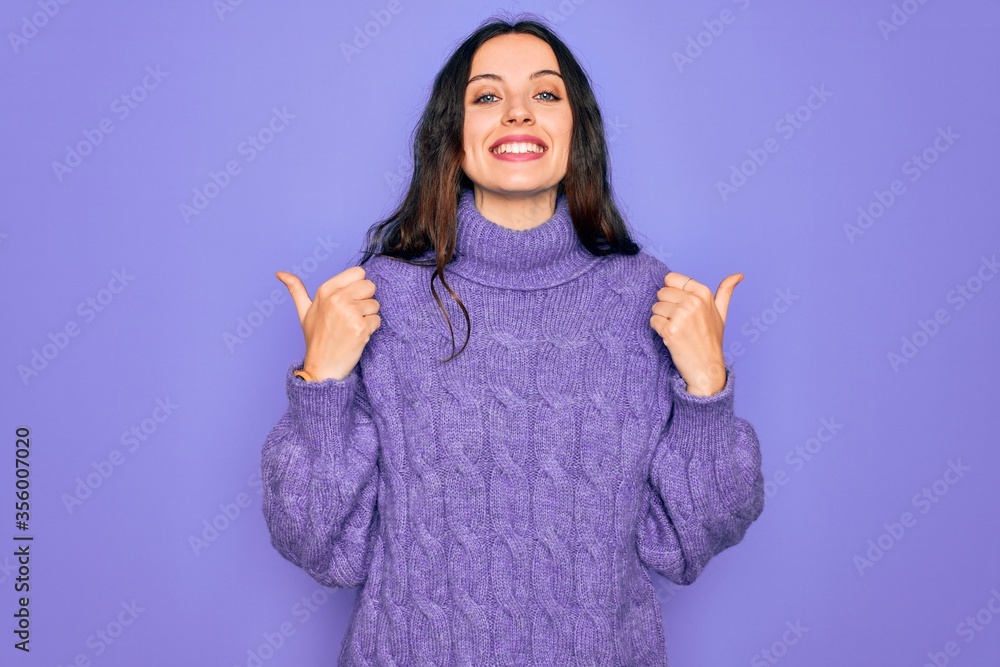 Wall mural Young beautiful woman wearing casual turtleneck sweater standing over purple background success sign doing positive gesture with hand, thumbs up smiling and happy. Cheerful expression and winner