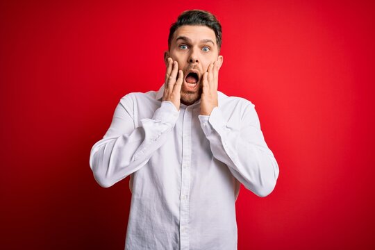 Young business man with blue eyes wearing elegant shirt standing over red isolated background afraid and shocked, surprise and amazed expression with hands on face