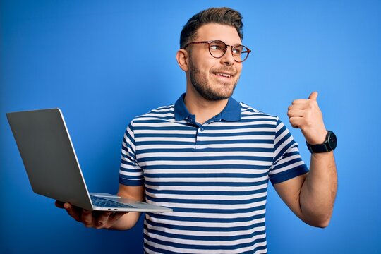 Young business man with blue eyes wearing glasses working using computer laptop pointing and showing with thumb up to the side with happy face smiling