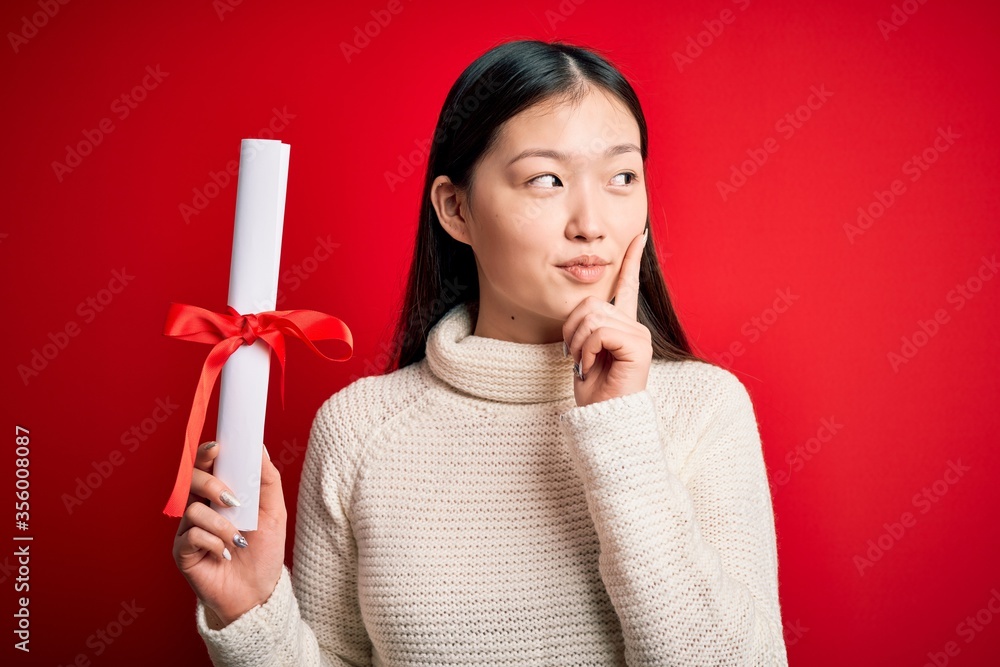 Canvas Prints Young asian student woman holding graduate diploma over red isolated background serious face thinking about question, very confused idea